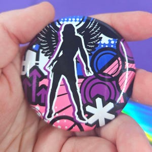 May include: A circular pin-back button featuring a black silhouette of a winged figure standing against a vibrant background of pink, purple, and blue geometric shapes, including arrows, circles, and a white star-like symbol with a glossy finish.