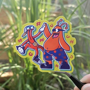 Puede incluir: Una pegatina con dos personajes de dibujos animados chocando los cinco. Uno es naranja y lleva gafas de sol y pantalones cortos azules con lunares. El otro es rojo y lleva un sombrero. La pegatina tiene un fondo amarillo con detalles verdes.