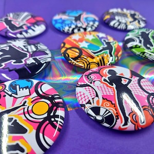 May include: A collection of circular pin-back buttons featuring vibrant, multi-colored graphic designs. Each button displays high-contrast black silhouettes of figures against abstract backgrounds of pink, yellow, orange, purple, and green patterns.