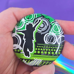 May include: A circular pinback button featuring a black silhouette of a person jumping with one arm raised. The background consists of a vibrant green, black, and white graphic design with circular patterns, dots, and geometric lines.