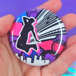 May include: A circular pinback button featuring a black silhouette of a person jumping against a vibrant background of purple, blue, and hot pink geometric shapes, circular patterns, and a white stylized city skyline silhouette at the bottom.