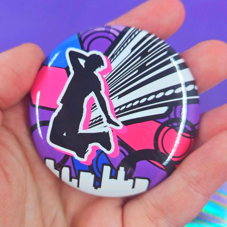 May include: A circular pinback button featuring a black silhouette of a person jumping against a vibrant background of purple, blue, and hot pink geometric shapes, circular patterns, and a white stylized city skyline silhouette at the bottom.