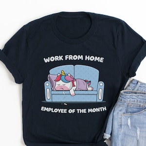 K&ouml;nnte beinhalten: Marineblaues T-Shirt mit einem Cartoon-Einhorn, das auf einem Sofa schl&auml;ft, mit dem Text "WORK FROM HOME EMPLOYEE OF THE MONTH". Das Shirt wird mit hellblauen Jeansshorts kombiniert.