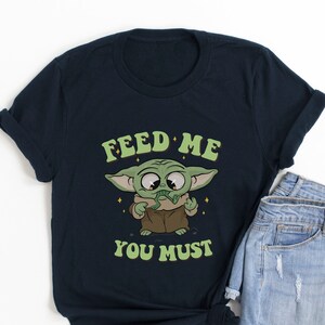 May include: Navy blue t-shirt featuring a cartoon character with the text "FEED ME YOU MUST". The character is green with large eyes and is holding a snack. The shirt is paired with denim shorts.