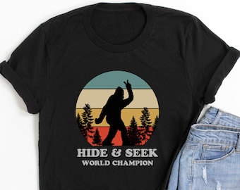May include: Black t-shirt featuring a Bigfoot silhouette giving a peace sign against a retro sunset design. The text "HIDE & SEEK WORLD CHAMPION" is printed below the graphic. The shirt is paired with denim shorts.