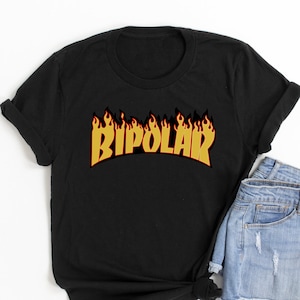 May include: Black t-shirt featuring the word "BIPOLAR" in yellow and orange flame-style lettering. The shirt is styled with denim shorts. The t-shirt is knotted at the bottom.
