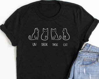 May include: Black t-shirt featuring a white outline design of four cats and the words "UN DEUX TROIS CAT". The shirt is styled with denim shorts.