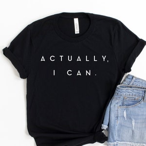 Op de afbeelding: Zwart t-shirt met de tekst "ACTUALLY, I CAN." in het wit, gecombineerd met lichtblauwe spijkerbroekshorts. Het t-shirt heeft opgerolde mouwen en is aan de zoom geknoopt. De shorts zijn versleten.