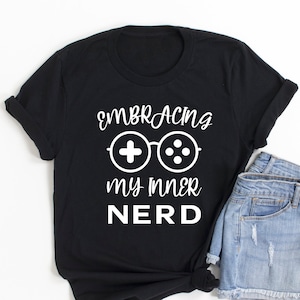 May include: Black t-shirt with the white text "Embracing my inner Nerd" and a controller graphic. The shirt is paired with denim shorts. The t-shirt is folded and tied at the bottom.