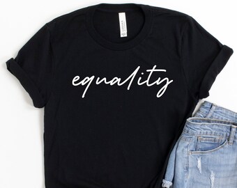 May include: Black t-shirt with the word "equality" in white script. The shirt is paired with light-wash denim shorts. The items are arranged on a white surface with abstract pink and blue shapes in the background.