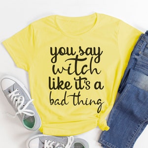 May include: A yellow t-shirt with the text "you say witch like it's a bad thing" in black script. The shirt is paired with blue jeans and gray sneakers with white laces, creating a casual outfit.