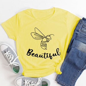 May include: A yellow t-shirt featuring a black line drawing of a bee and the word "Beautiful". The image also shows blue jeans and grey trainers with white laces, a casual outfit.
