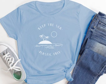 May include: A light blue t-shirt with the text "KEEP THE SEA. PLASTIC FREE." is paired with blue jeans and gray sneakers. The shirt features a white graphic of an island with palm trees, a sun, and ocean waves.