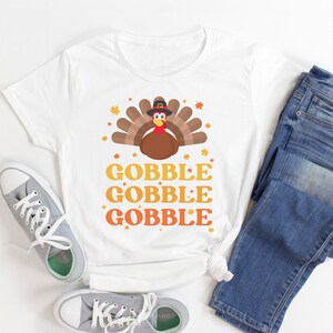 May include: White t-shirt with a Thanksgiving turkey graphic and the words "GOBBLE GOBBLE GOBBLE" in orange and yellow. The shirt is paired with blue jeans and gray sneakers.