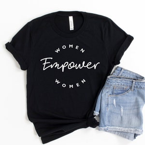 May include: Black t-shirt with the text "WOMEN Empower WOMEN" in white script. The shirt is paired with light-wash denim shorts. The shirt is folded at the sleeves and knotted at the bottom.