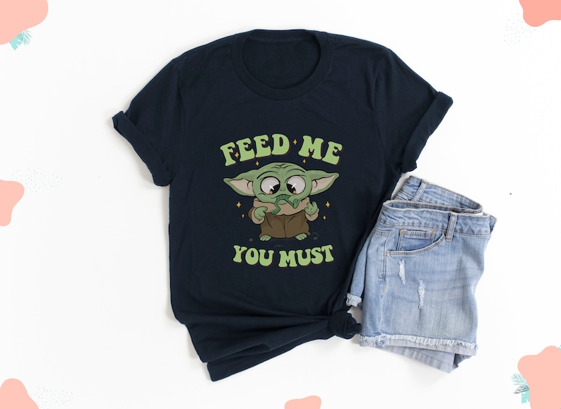 May include: Navy blue t-shirt featuring a cartoon character with the text "FEED ME YOU MUST". The character is green with large eyes and is holding a snack. The shirt is paired with denim shorts.