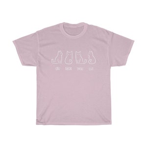 May include: A light pink t-shirt featuring a white line drawing of four cats. The text "UN DEUX TROIS CAT" is printed below the cats. The shirt is made of a soft material.