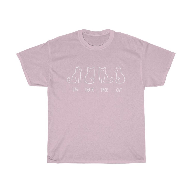May include: A light pink t-shirt featuring a white line drawing of four cats. The text "UN DEUX TROIS CAT" is printed below the cats. The shirt is made of a soft material.