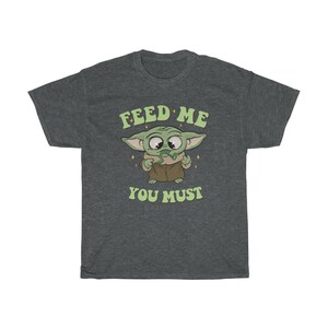 May include: Dark gray t-shirt featuring a cartoon character with large eyes and green skin. The character is holding a green object and wearing a brown robe. The text "FEED ME YOU MUST" is printed above and below the character in a retro font.