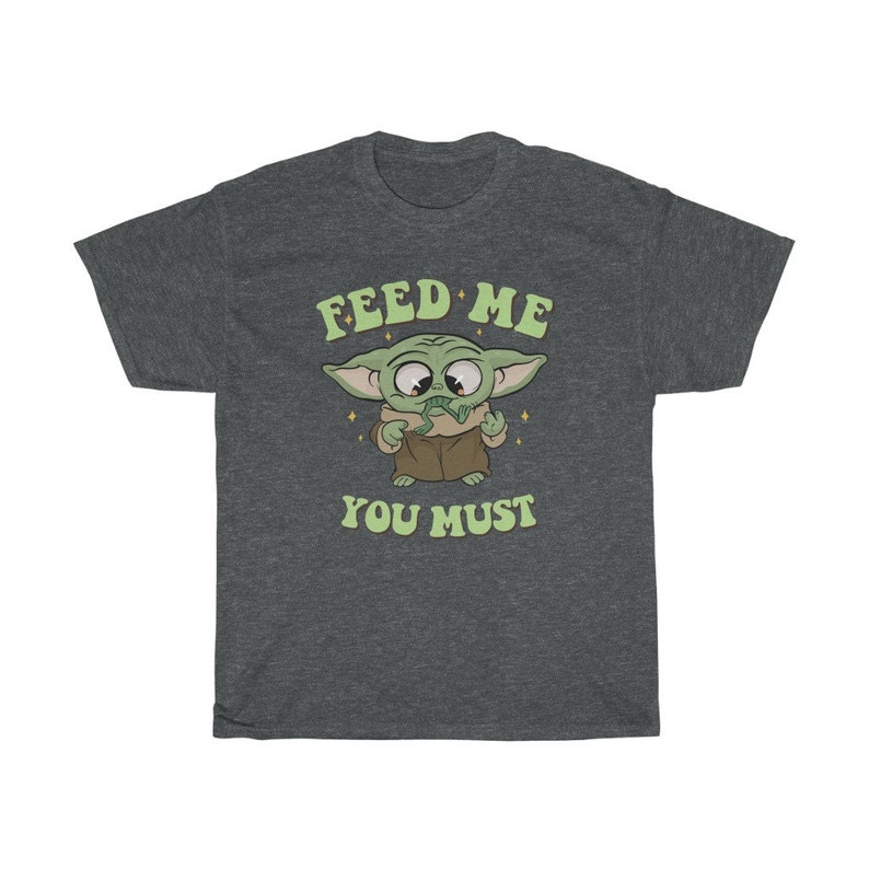 May include: Dark gray t-shirt featuring a cartoon character with large eyes and green skin. The character is holding a green object and wearing a brown robe. The text "FEED ME YOU MUST" is printed above and below the character in a retro font.