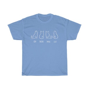 May include: Light blue t-shirt featuring a playful design of four white cat illustrations. The text "UN DEUX TROIS CAT" is printed below the cats. A casual and comfortable tee.
