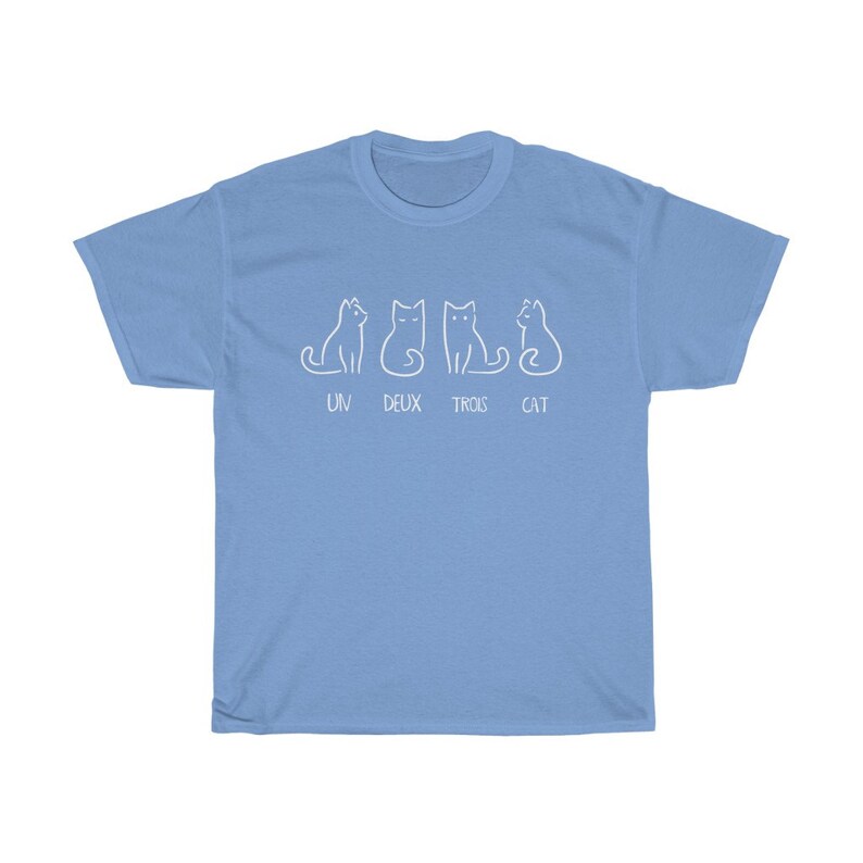 May include: Light blue t-shirt featuring a playful design of four white cat illustrations. The text "UN DEUX TROIS CAT" is printed below the cats. A casual and comfortable tee.