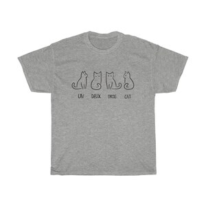 May include: A heather gray t-shirt featuring a minimalist design of four black cat illustrations. The text "UN DEUX TROIS CAT" is printed below the cats. The shirt is made of a soft, comfortable material.