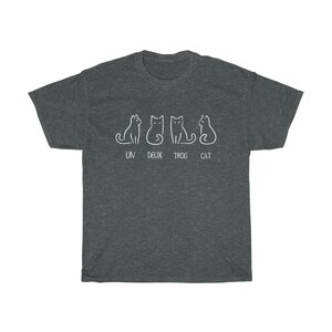 May include: Dark gray t-shirt featuring a white line drawing of four cats in different poses. Below the cats, the text reads "UN DEUX TROIS CAT". The shirt is made of a soft, comfortable material.