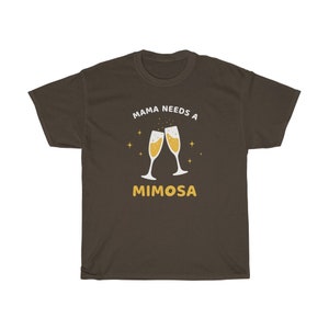 May include: A dark brown t-shirt with the text "MAMA NEEDS A MIMOSA" in white and yellow. The design features two champagne flutes with bubbly liquid and star accents.