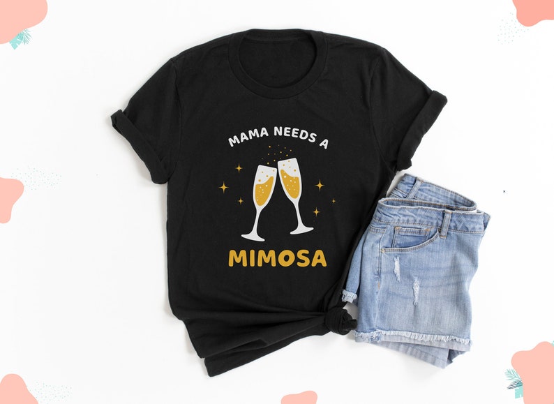 May include: Black t-shirt with the text "MAMA NEEDS A MIMOSA" in white and yellow. The design features two champagne flutes with bubbly liquid, and gold stars. The shirt is paired with denim shorts.