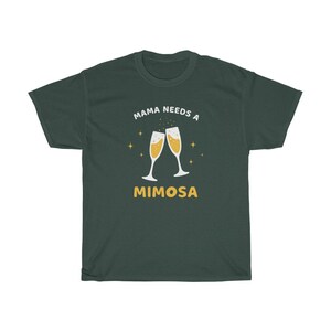 May include: Dark green t-shirt with the text "MAMA NEEDS A MIMOSA" in white and yellow. The design features two champagne flutes with bubbly liquid and star accents. A casual, comfortable tee.