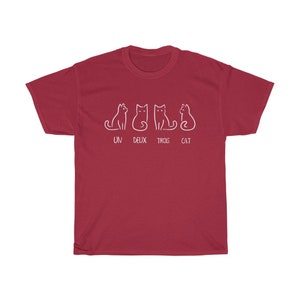 May include: A maroon t-shirt featuring a white line drawing of four cats in different poses. Below the cats, the words "UN DEUX TROIS CAT" are printed in white. The shirt is made of a soft, comfortable material.