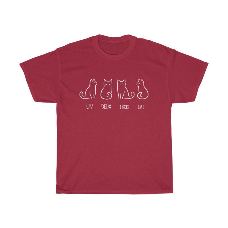 May include: A maroon t-shirt featuring a white line drawing of four cats in different poses. Below the cats, the words "UN DEUX TROIS CAT" are printed in white. The shirt is made of a soft, comfortable material.