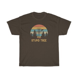 Pode incluir: Camiseta marrom com um design retr&ocirc; de disc golf. O gr&aacute;fico mostra uma cesta de disc golf, um disco e &aacute;rvores contra um p&ocirc;r do sol. O texto "STUPID TREE" est&aacute; abaixo. Para os entusiastas do disc golf.
