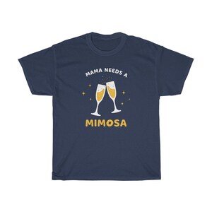 May include: Navy blue t-shirt with the text "MAMA NEEDS A MIMOSA" in white and yellow. The design features two champagne flutes with bubbly liquid and star accents. A casual, comfortable tee.