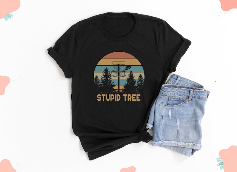 Pode incluir: Camiseta preta com um design de disc golf. O gr&aacute;fico inclui um p&ocirc;r do sol, &aacute;rvores, uma cesta de disc golf e o texto "STUPID TREE" em uma fonte de estilo vintage. A camiseta &eacute; combinada com shorts jeans.