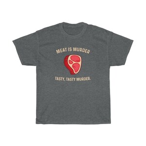 Op de afbeelding: Donkergrijs t-shirt met de tekst "MEAT IS MURDER" boven een cartoon steak illustratie. Onder de illustratie staat de tekst "TASTY, TASTY MURDER".