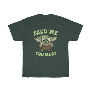 May include: Dark green t-shirt featuring a cartoon character with large eyes and green skin. The character is holding a green object and the text "FEED ME YOU MUST" is above and below the character in a retro font.