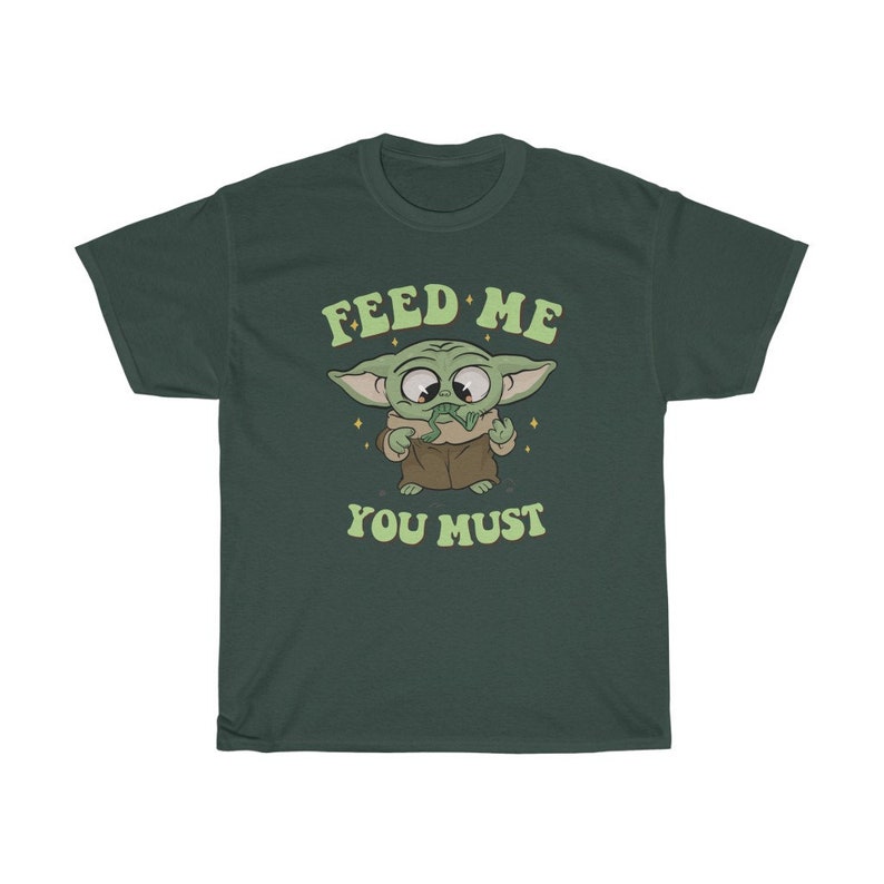 May include: Dark green t-shirt featuring a cartoon character with large eyes and green skin. The character is holding a green object and the text "FEED ME YOU MUST" is above and below the character in a retro font.
