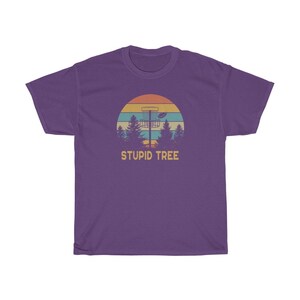 Pode incluir: Camiseta roxa com um design retr&ocirc; com uma cesta de disc golf, &aacute;rvores e um p&ocirc;r do sol. O texto "STUPID TREE" &eacute; impresso abaixo. Uma camiseta casual para os entusiastas do disc golf.