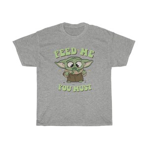 May include: A heather gray t-shirt featuring a cartoon image of a green alien character with large eyes and pointed ears. The text "FEED ME YOU MUST" is printed above and below the character in green lettering.