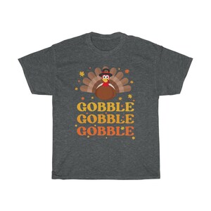 May include: Dark gray t-shirt featuring a cartoon turkey wearing a pilgrim hat, with the words "GOBBLE GOBBLE GOBBLE" in orange and yellow. The design includes fall leaves and stars, perfect for Thanksgiving.