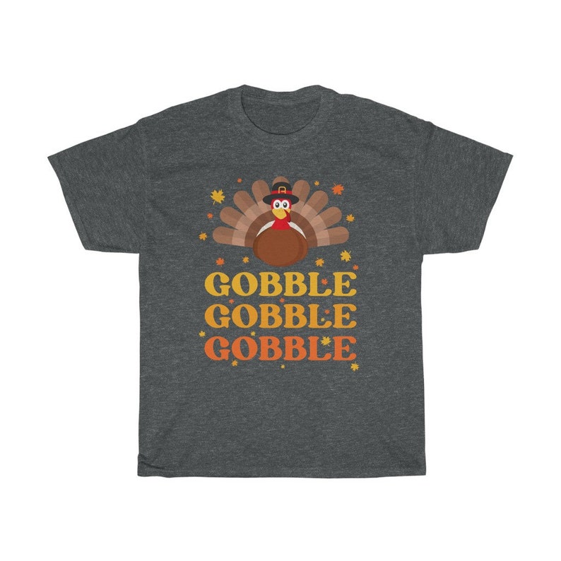 May include: Dark gray t-shirt featuring a cartoon turkey wearing a pilgrim hat, with the words "GOBBLE GOBBLE GOBBLE" in orange and yellow. The design includes fall leaves and stars, perfect for Thanksgiving.