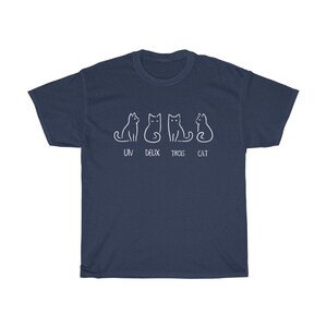 May include: Navy blue t-shirt featuring a white graphic of four stylized cats in different poses. Below the cats, the text reads "UN DEUX TROIS CAT". The shirt is a casual wear item.
