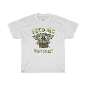 May include: White t-shirt featuring a cartoon character with large eyes and green skin, wearing a brown robe. The text "FEED ME YOU MUST" is printed above and below the character in green, retro-style lettering.