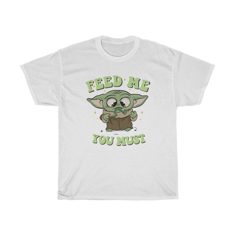May include: White t-shirt featuring a cartoon character with large eyes and green skin, wearing a brown robe. The text "FEED ME YOU MUST" is printed above and below the character in green, retro-style lettering.