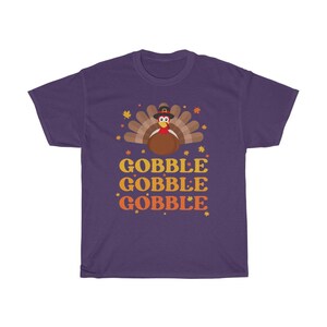 May include: A purple t-shirt featuring a cartoon turkey wearing a pilgrim hat, with the words "GOBBLE GOBBLE GOBBLE" in orange and yellow. The design includes fall leaves and stars, perfect for Thanksgiving.