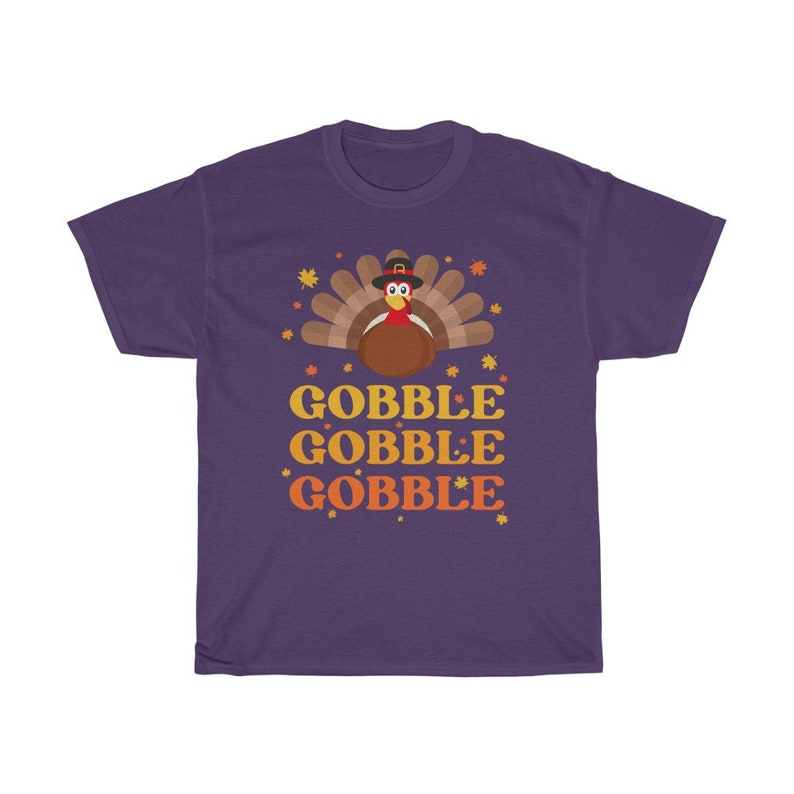 May include: A purple t-shirt featuring a cartoon turkey wearing a pilgrim hat, with the words "GOBBLE GOBBLE GOBBLE" in orange and yellow. The design includes fall leaves and stars, perfect for Thanksgiving.