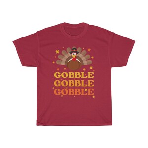 May include: A maroon t-shirt featuring a cartoon turkey wearing a pilgrim hat, with a fan of brown and tan feathers. The text "GOBBLE GOBBLE GOBBLE" is printed in orange and yellow below the turkey illustration. Autumn leaves are scattered around the design.