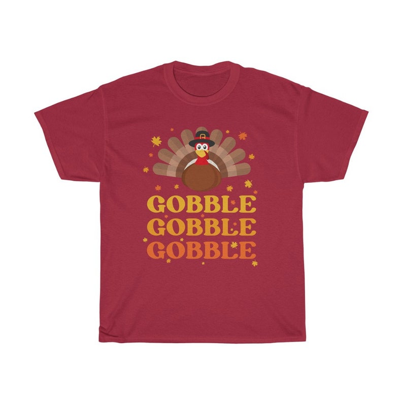 May include: A maroon t-shirt featuring a cartoon turkey wearing a pilgrim hat, with a fan of brown and tan feathers. The text "GOBBLE GOBBLE GOBBLE" is printed in orange and yellow below the turkey illustration. Autumn leaves are scattered around the design.